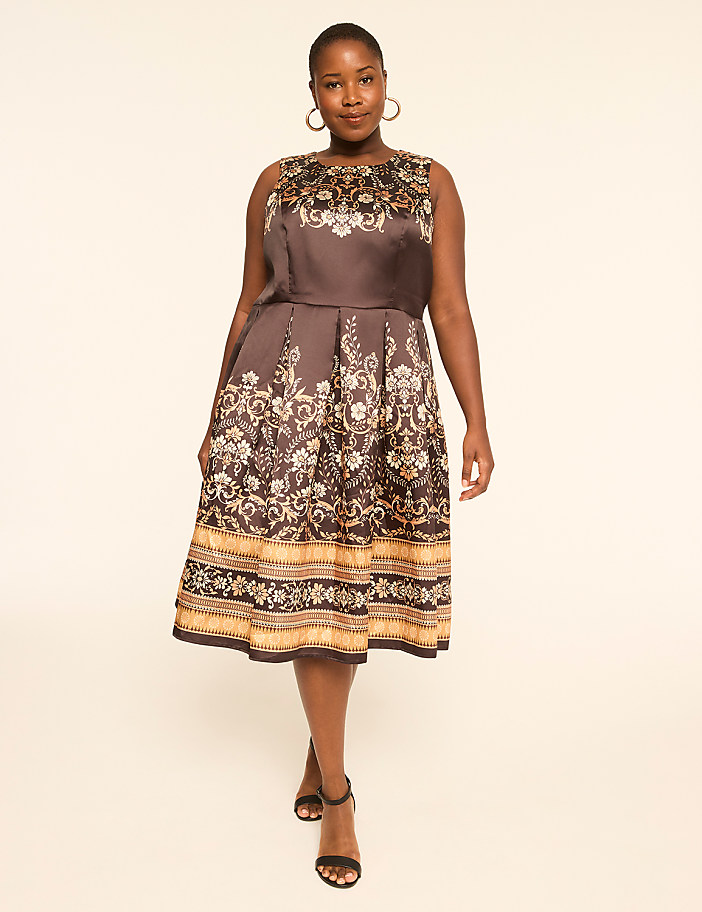 A woman in a patterned dress stands against a plain, light-colored background.