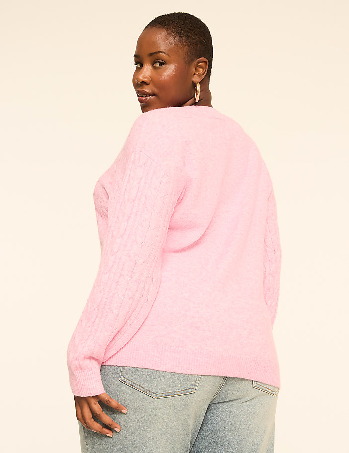 A woman in a pink cable-knit sweater and light-wash jeans poses against a plain, light-colored background.