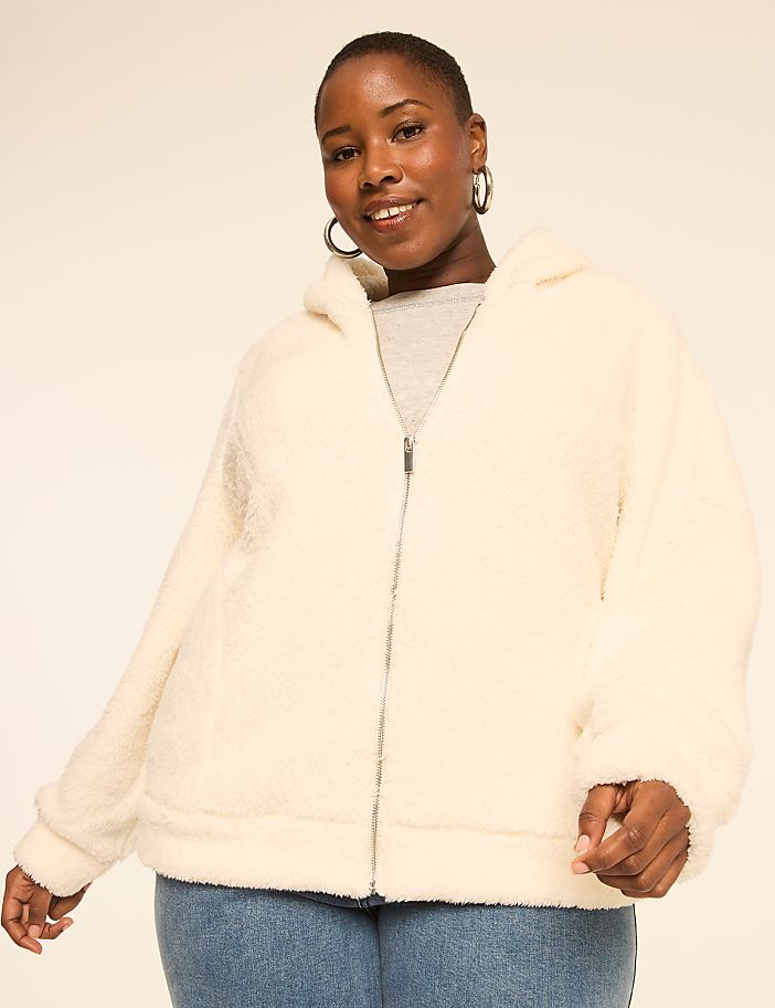 A woman in a cream-colored fuzzy jacket and blue jeans smiles against a plain, light background.