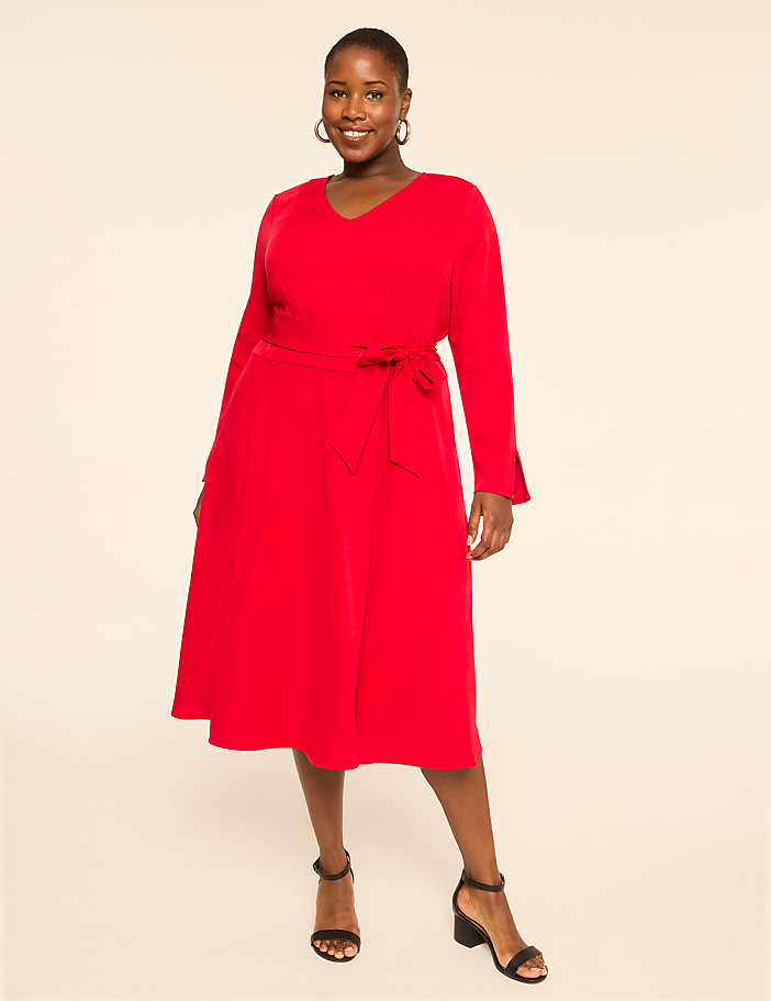 A smiling woman in a red dress stands against a plain, light-colored background.