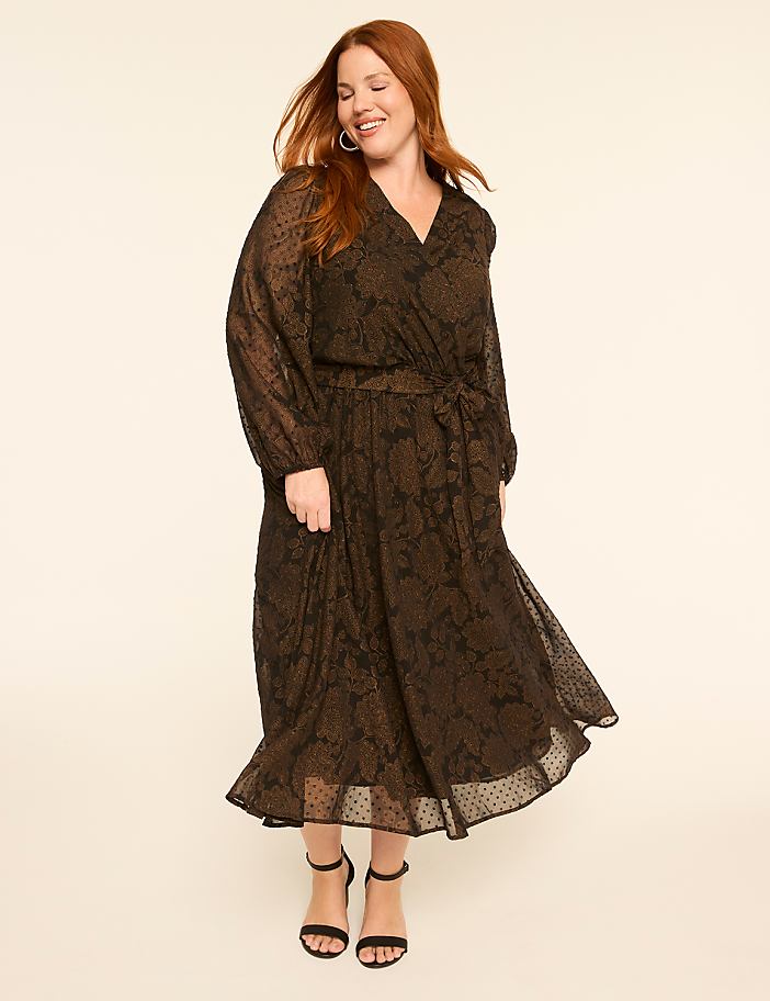 A smiling woman with red hair in a long, patterned black dress stands against a plain, light-colored background.