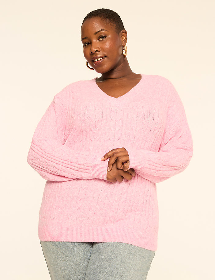 A woman in a pink cable-knit sweater stands against a plain, light-colored background.