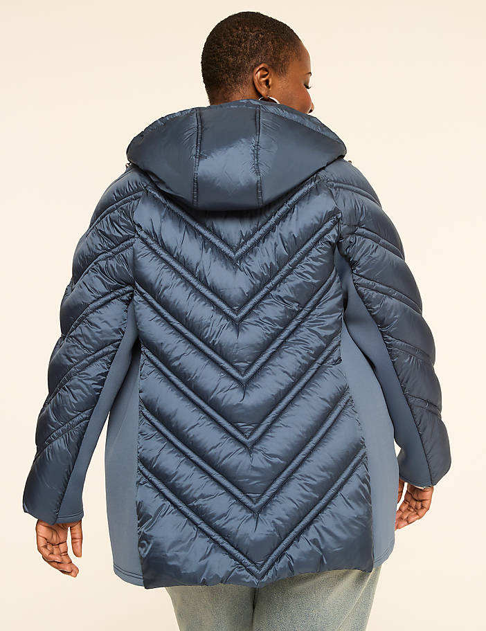 A woman in a blue puffer jacket with a chevron pattern on the back stands against a plain, light-colored background.