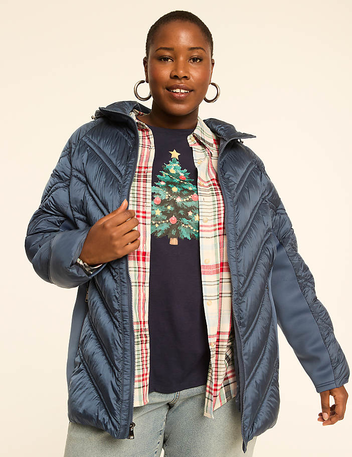 A woman in a blue puffer jacket and a Christmas tree t-shirt smiles against a plain background.