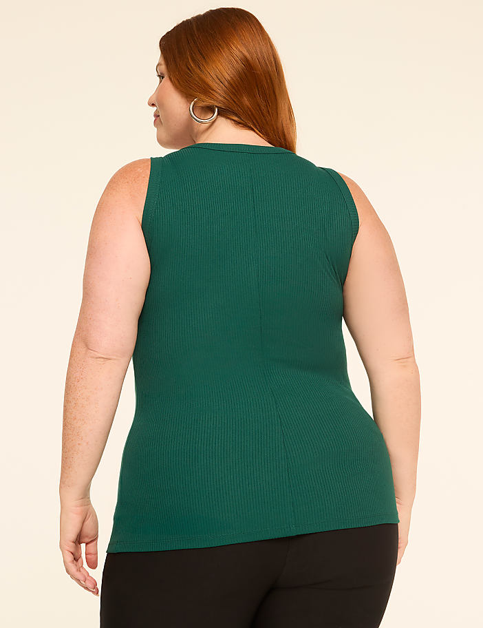 A woman with red hair wearing a green ribbed tank top and black pants stands against a plain beige background.