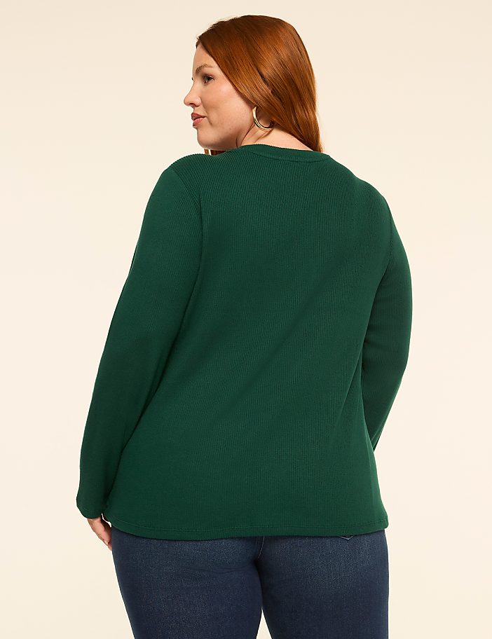 A woman with red hair, wearing a dark green long-sleeved shirt and blue jeans, stands against a plain, light-colored background.