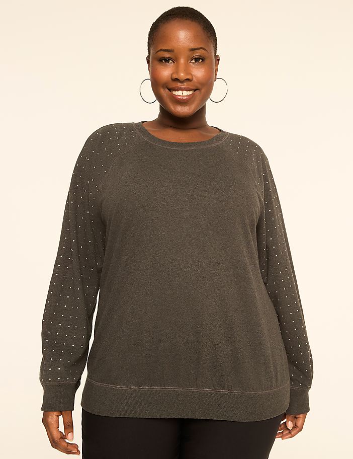 A smiling woman in a dark grey, studded sweater stands against a plain, light background.