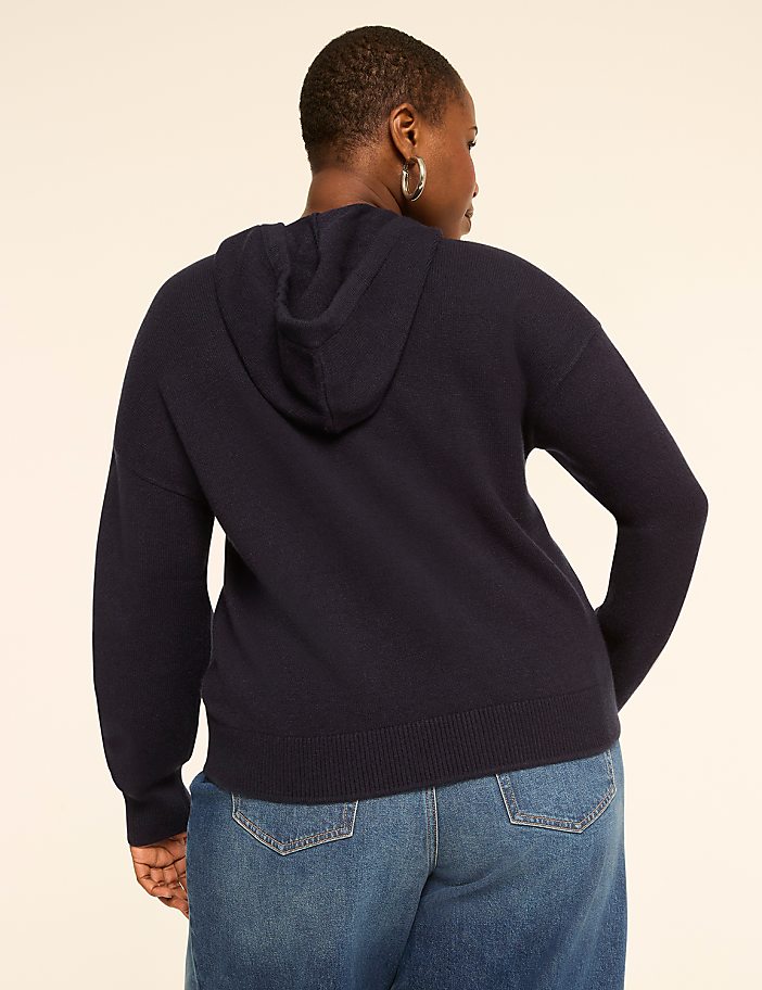 A woman in a navy hoodie and jeans stands against a plain beige background.