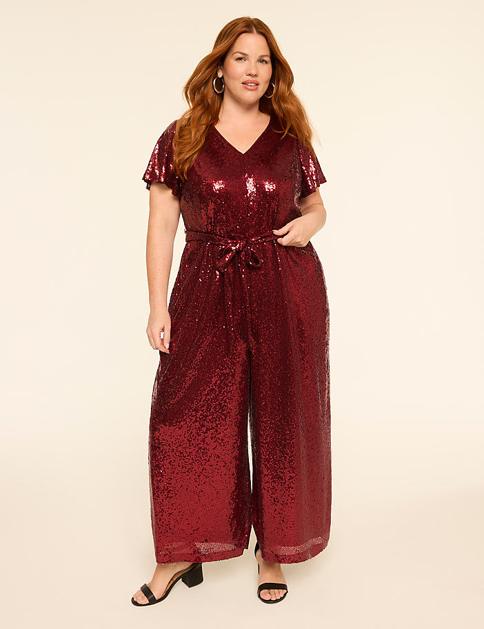 A woman in a sparkling red jumpsuit stands against a plain, light-colored background.