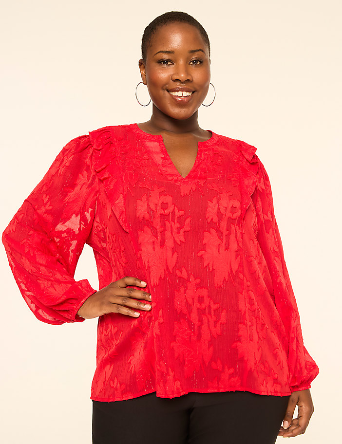 A smiling woman in a red textured blouse stands against a plain, light-colored background.
