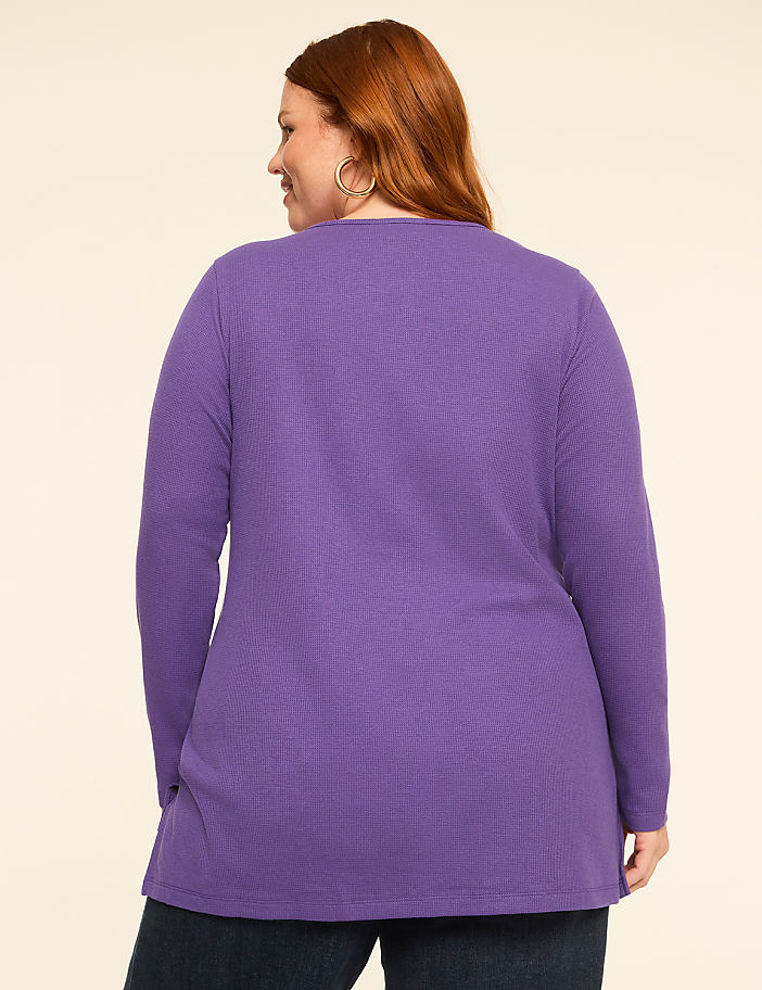 A woman with red hair wearing a purple long-sleeved shirt stands against a plain, light-colored background.