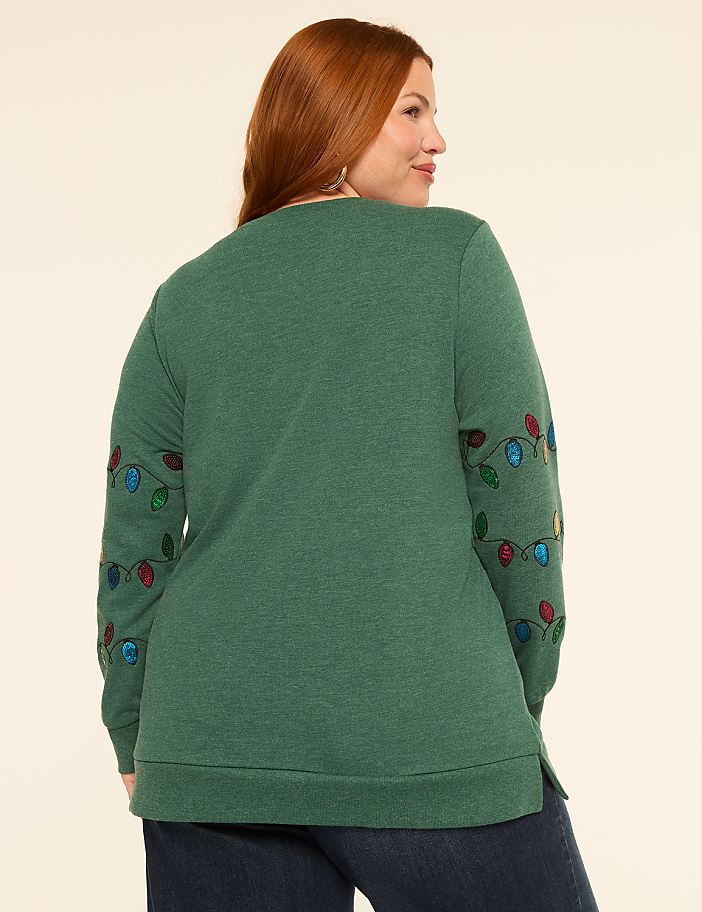 A woman with red hair wearing a green sweatshirt with Christmas lights on the sleeves stands against a plain, light-colored background.