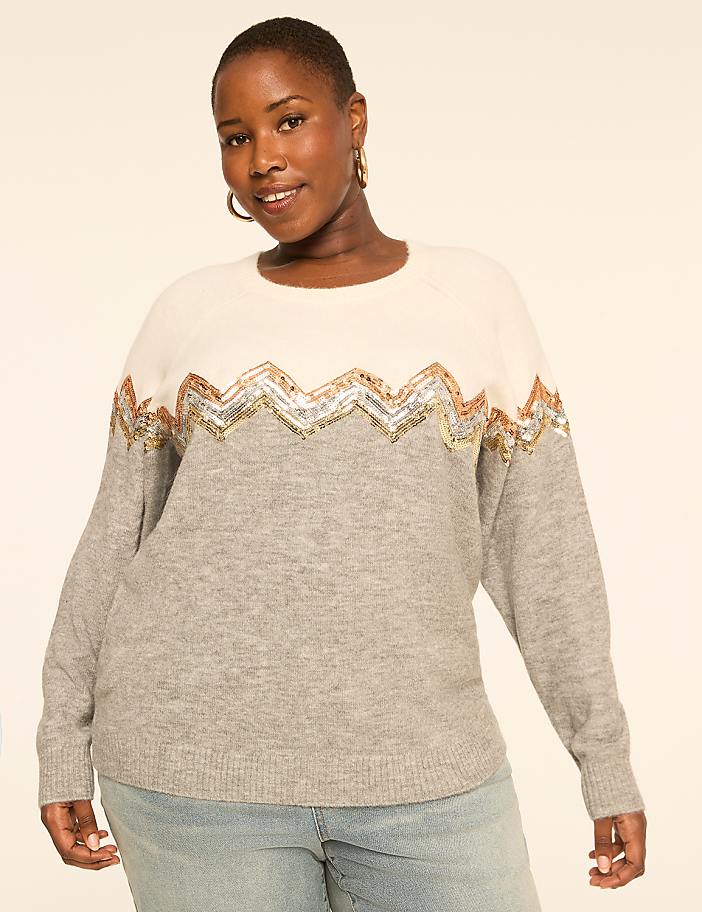 A woman in a light grey and cream sweater with a sequined zigzag pattern stands against a plain, light-colored background.