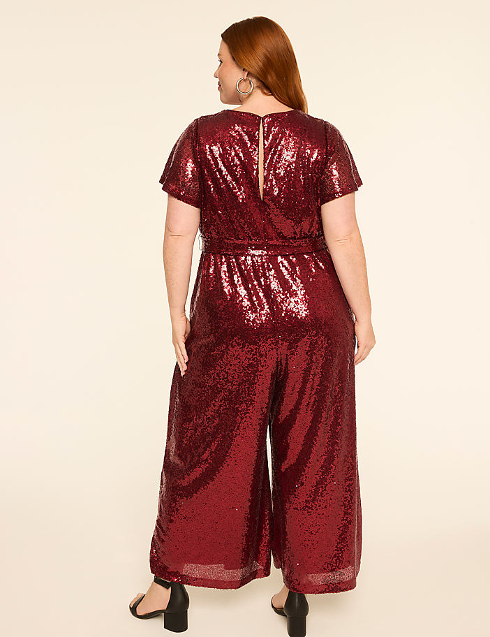 A woman in a sparkly red jumpsuit stands against a plain, light-colored background.