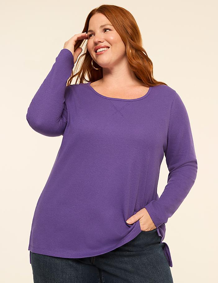A smiling woman with red hair in a purple long-sleeved shirt and jeans poses against a plain, light-colored background.