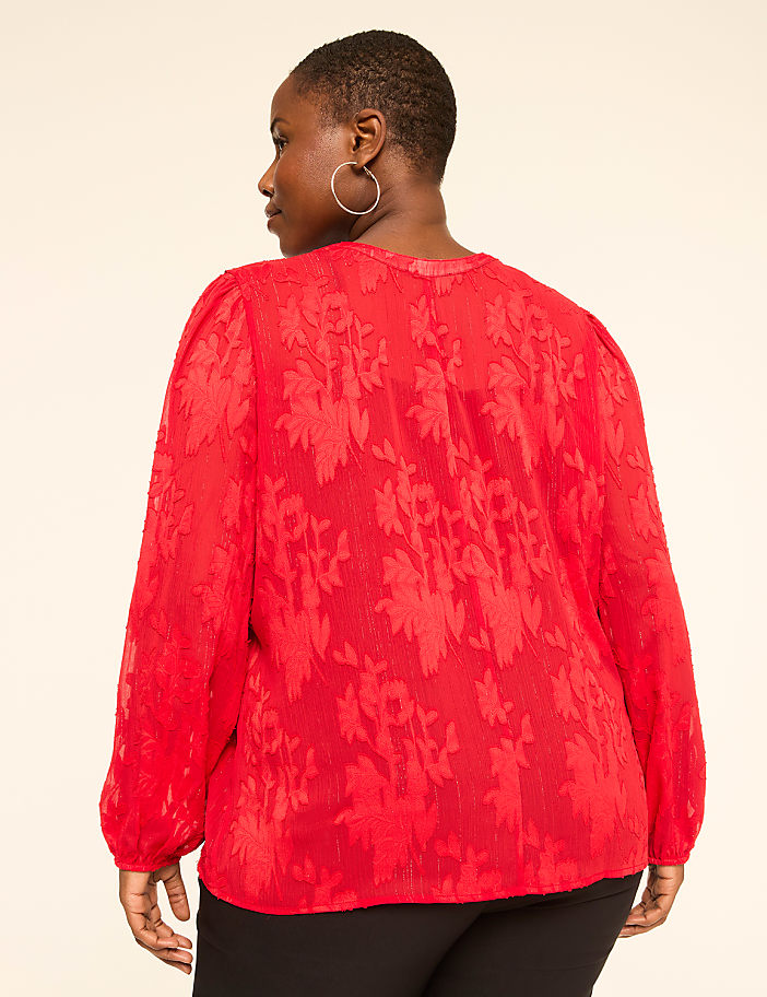A woman in a red floral textured blouse stands against a plain, light-colored background.