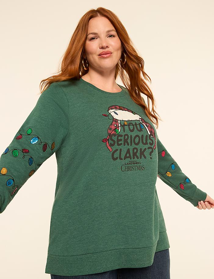 A woman with red hair smiles at the camera wearing a green Christmas sweater with a "You Serious, Clark?" graphic and colorful lights on the sleeves, against a plain, light-colored background.
