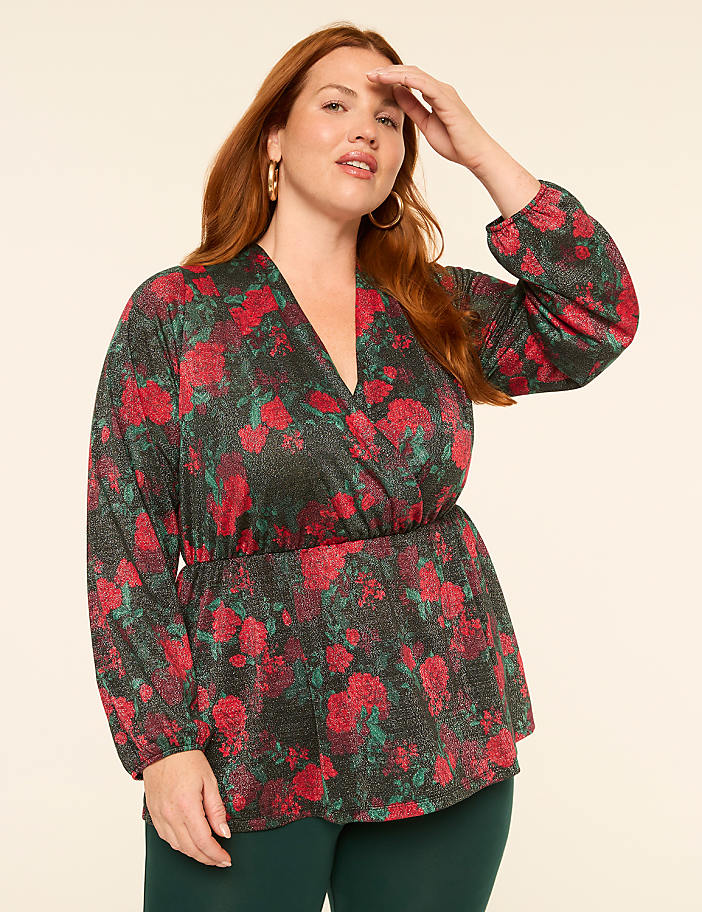 A woman with red hair poses in a sparkly floral wrap top and green pants against a plain cream background.