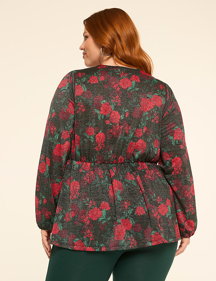 A woman with red hair, wearing a long-sleeved floral peplum top and green leggings, stands against a plain, light-colored background.