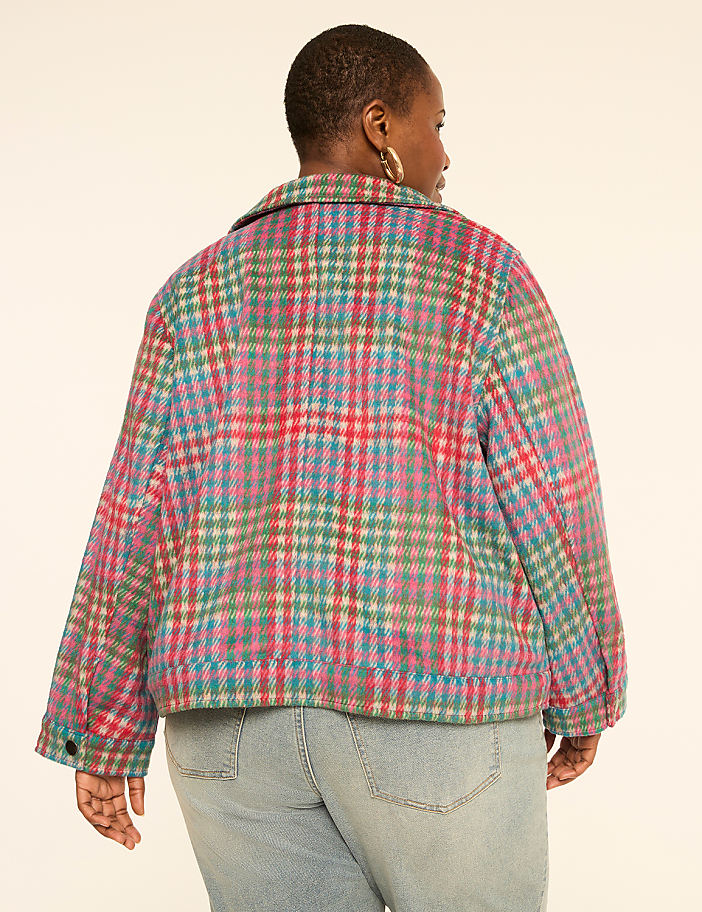 A woman wearing a colorful plaid jacket and light blue jeans stands against a plain, light-colored background.
