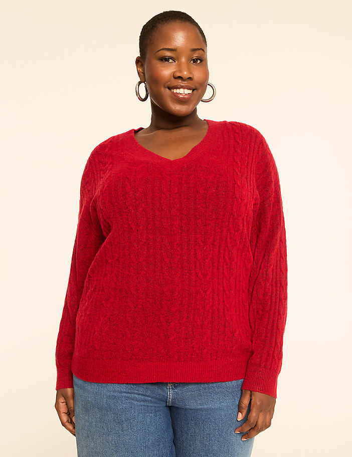 A smiling woman in a red cable-knit sweater and blue jeans stands against a plain, light-colored background.