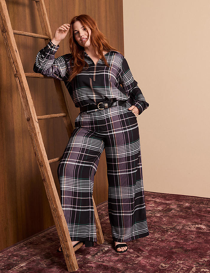 A woman in a plaid outfit leans against a wooden ladder in front of a warm-toned wall and a patterned rug.