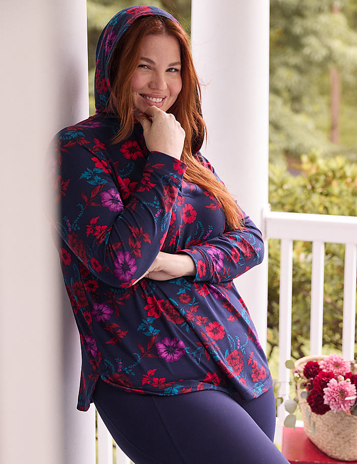 A smiling woman in a floral hoodie leans against a white pillar on a porch with a basket of flowers in the background.
