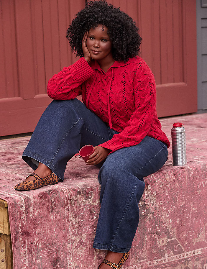 A woman in a red cable-knit sweater and wide-leg jeans sits on a patterned rug with a thermos and mug, in front of a red wooden wall.