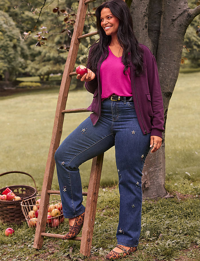 A woman in jeans and a cardigan stands by a ladder with apples in a basket in the foreground, while a tree and grassy yard form the background.