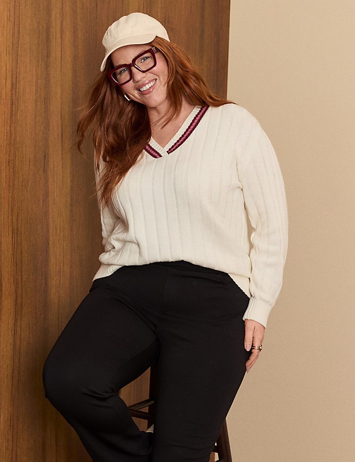 A smiling woman with red hair, wearing a cream sweater, black pants, and a baseball cap, poses in front of a wood-paneled wall.