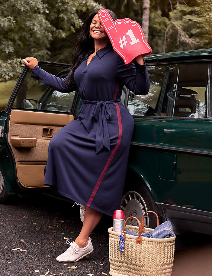 A woman in a navy dress and white sneakers holds a red foam finger while leaning against a vintage car, with a picnic basket and thermos in the foreground and trees in the background.