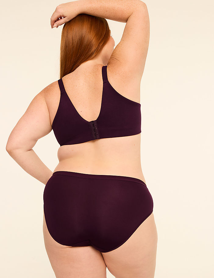 A woman with auburn hair stands with her back to the viewer, wearing a plum-colored bra and matching underwear against a plain, light-colored background.