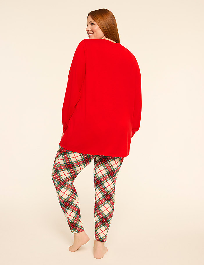 A woman in a red sweater and plaid pajama bottoms stands against a plain, light-colored background.