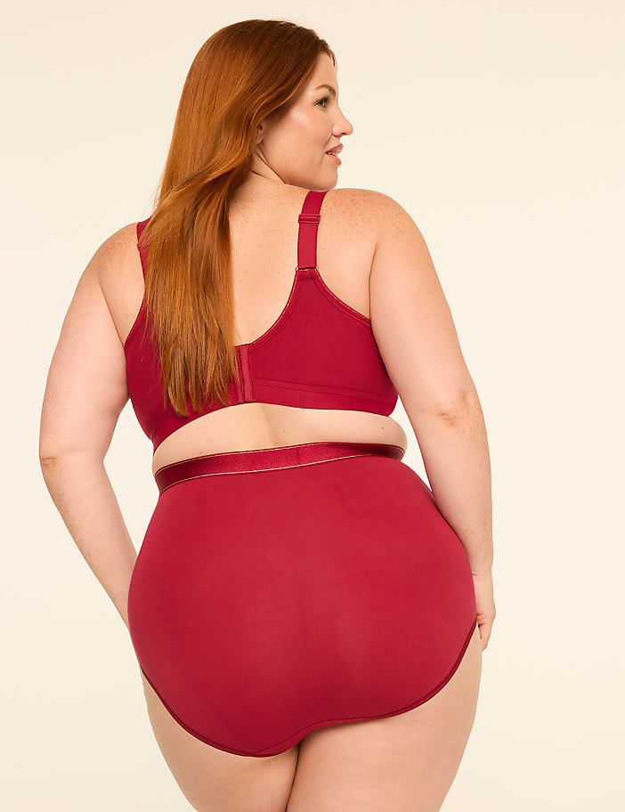A woman with red hair, wearing a red bra and high-waisted underwear, stands against a plain, light-colored background.