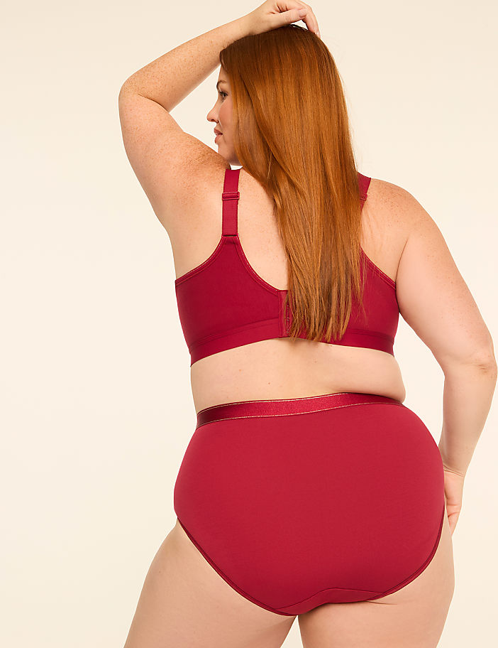 A woman with red hair in a maroon bra and underwear stands against a plain, light background.