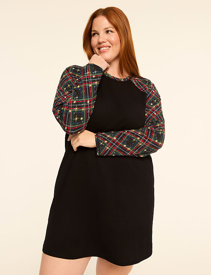 A smiling woman with red hair poses in a black dress with plaid sleeves against a plain, light-colored background.