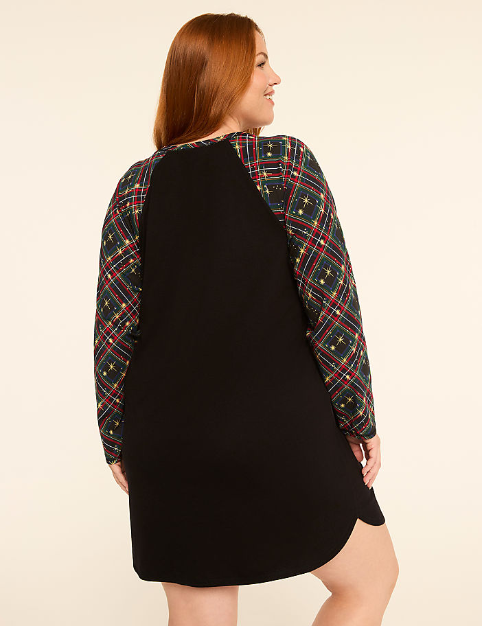 A woman with red hair stands facing away from the camera, wearing a black dress with plaid sleeves, against a plain, light-colored background.