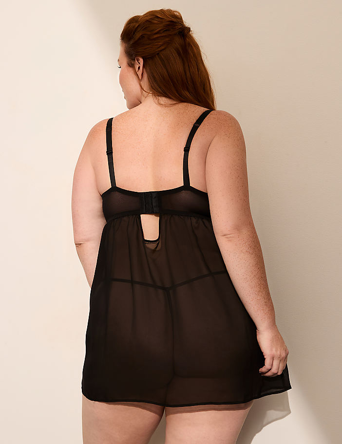 A woman with red hair stands facing away from the camera in a sheer black negligee against a plain, light-colored background.