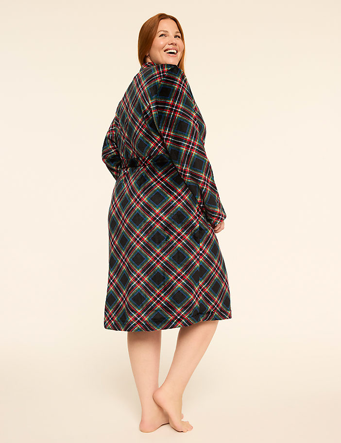 A smiling woman in a plaid robe stands barefoot against a plain, light-colored background.