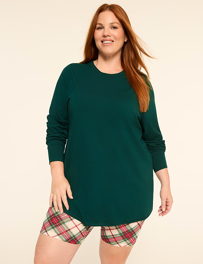 A smiling woman with red hair stands in the foreground wearing a green long-sleeved shirt and plaid shorts against a plain, light-colored background.