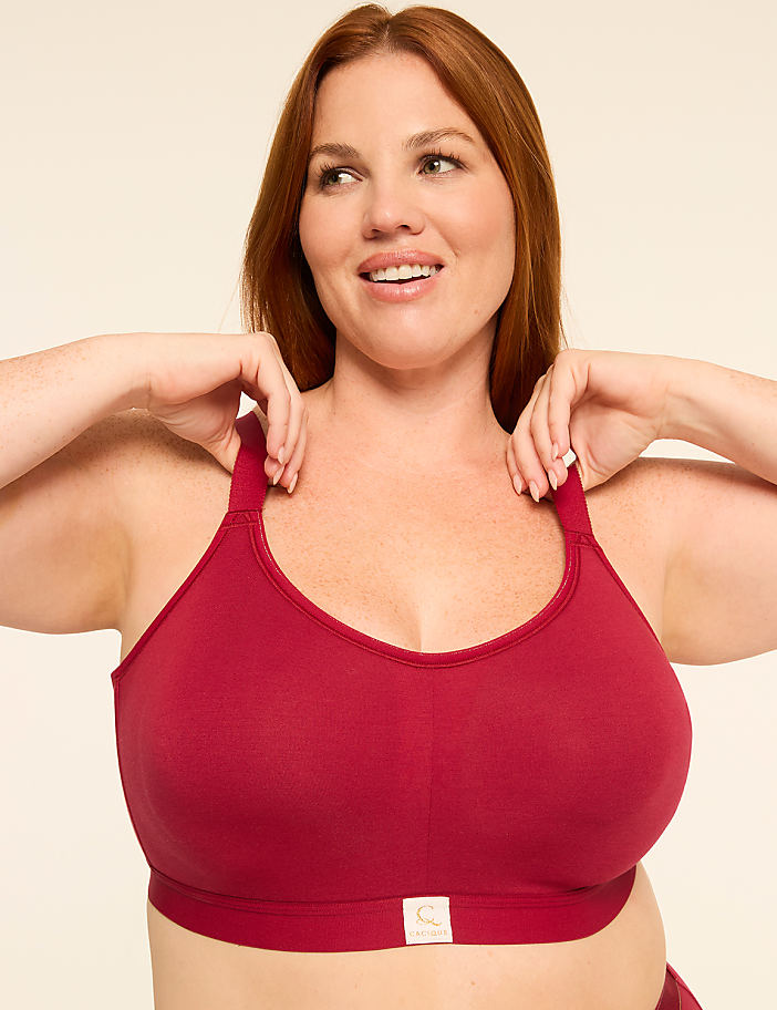 A smiling woman with red hair in a maroon sports bra adjusts the straps against a plain cream background.