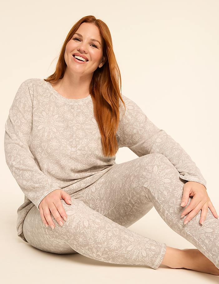 A smiling woman with red hair sits on a light-colored floor wearing a matching patterned loungewear set, with a plain, light background behind her.