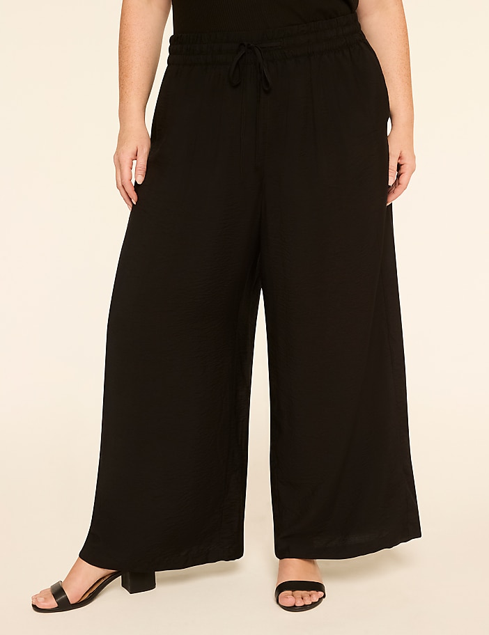 A person wearing black wide-leg pants and sandals stands against a plain, light-colored background.