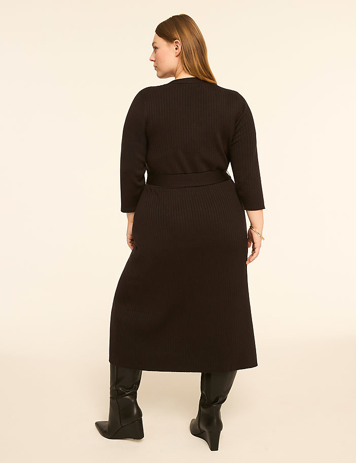A woman in a black ribbed dress and boots stands against a plain, light-colored background.