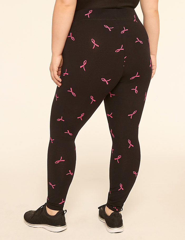 A person wearing black leggings with a pink ribbon pattern stands against a plain, light-colored background.