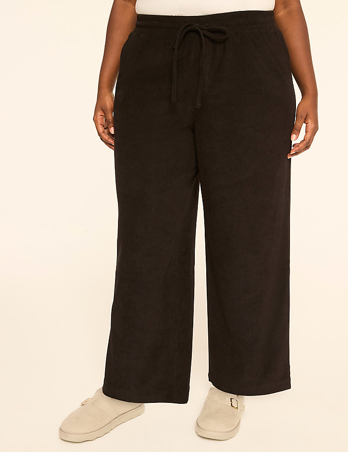 A person wearing black wide-leg sweatpants and light-colored slippers stands against a plain, light-colored background.