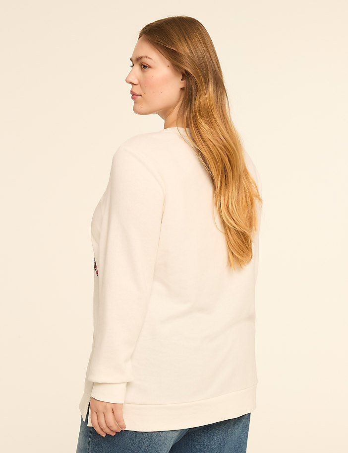 A woman in a cream sweatshirt and blue jeans stands against a plain, light-colored background.