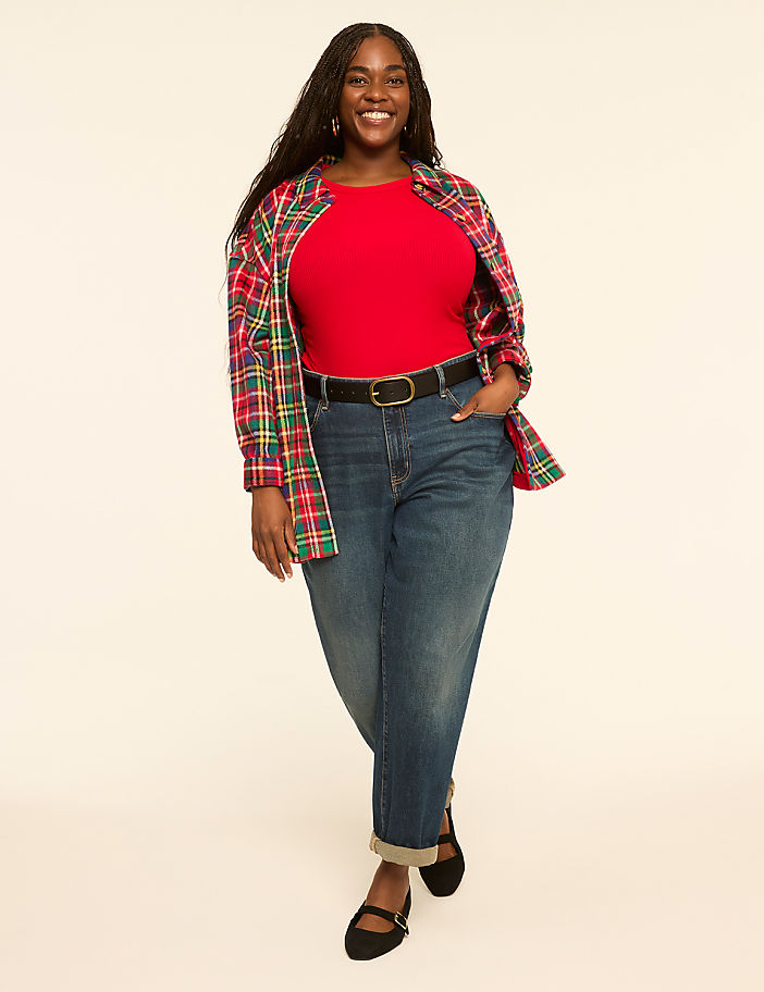 A smiling woman in a red shirt, jeans, and a plaid jacket stands against a plain, light-colored background.