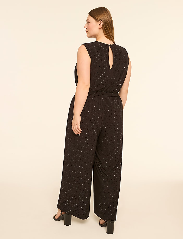 A woman in a black polka dot jumpsuit stands against a plain, light-colored background.