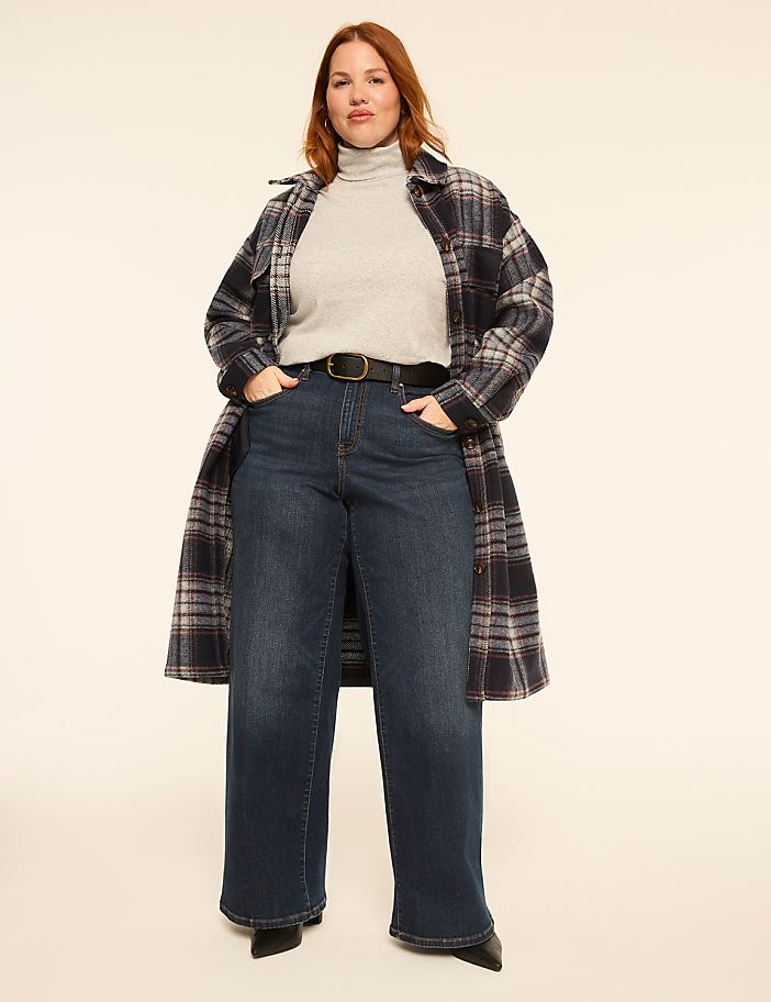 A woman in a plaid coat, turtleneck, and wide-leg jeans stands against a plain, light-colored background.